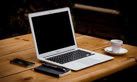 Ordinateur portable ouvert sur une table en bois, écran entièrement noir, à côté d’une tasse de café et d’un carnet fermé.